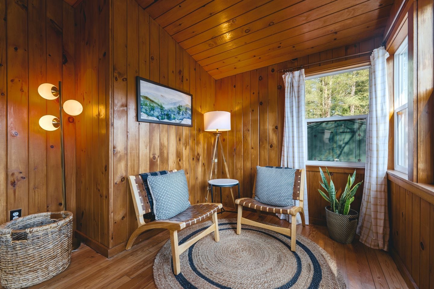 A wooden cabin interior with two chairs, a lamp, a rug, and a window with curtains.