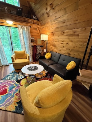 A cozy room with a wooden ceiling, a sofa, a yellow armchair, a small table, and a colorful rug.