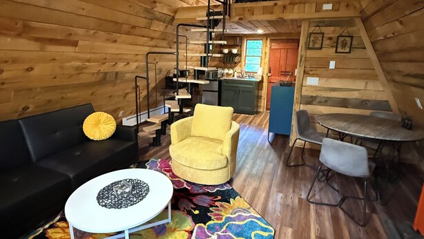 L’intérieur d’un chalet accueillant, avec un plafond en bois, un canapé, un fauteuil jaune, une table à manger et un escalier menant à un niveau supérieur.