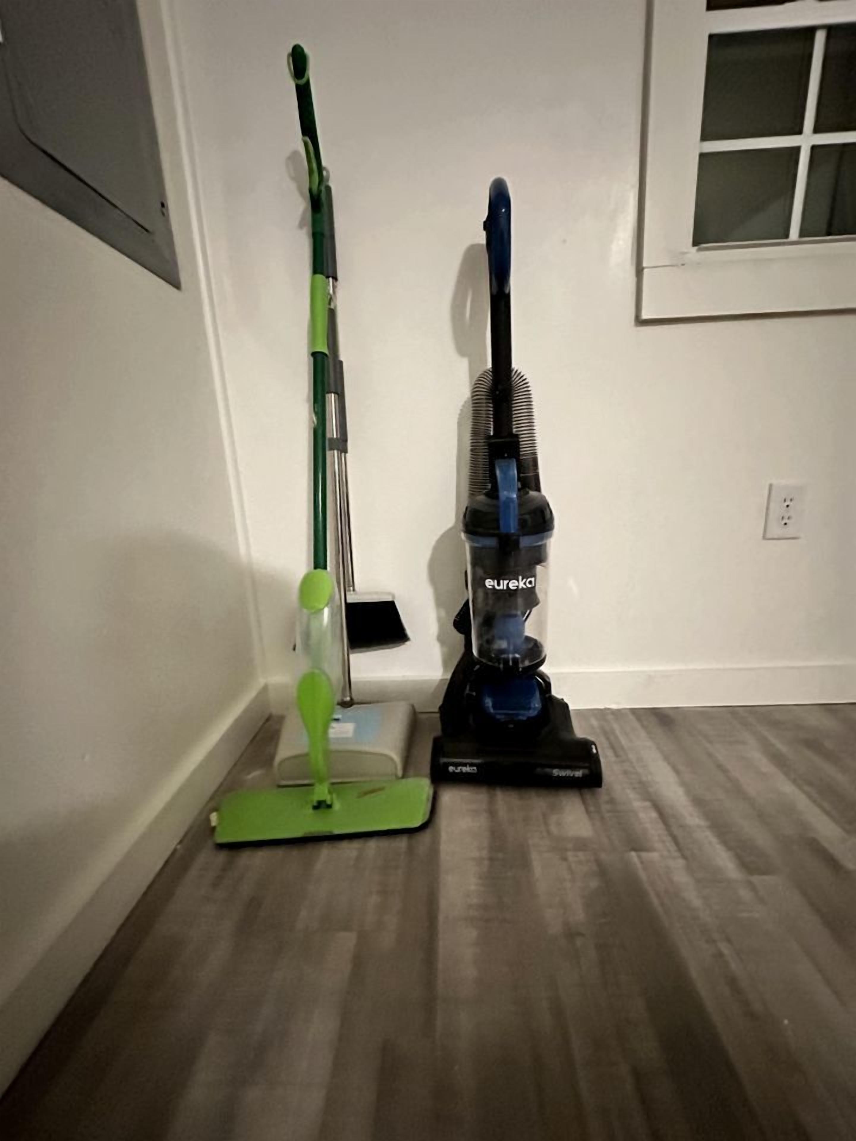 Ein Zimmer mit Holzboden, einem grün-schwarz gestreiften Mop und einem blau-schwarz gefärbten Staubsauger.