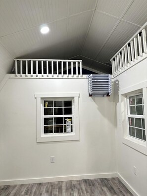 Une pièce avec un plafond en pente, une fenêtre avec des stores, un radiateur et une rambarde blanche sur le haut du mur.