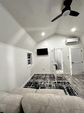Une pièce dotée d’un ventilateur de plafond, d’un téléviseur fixé au mur, d’un tapis à motifs et d’une porte donnant sur une autre pièce.