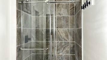 A glass-enclosed shower stall with a white bathtub, marble tiles, and a handrail.