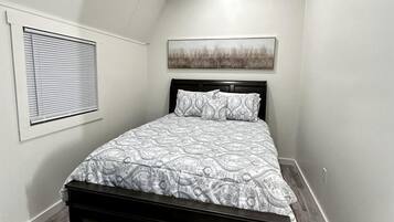A bedroom with a bed, a window with blinds, a painting on the wall, and a ceiling light.