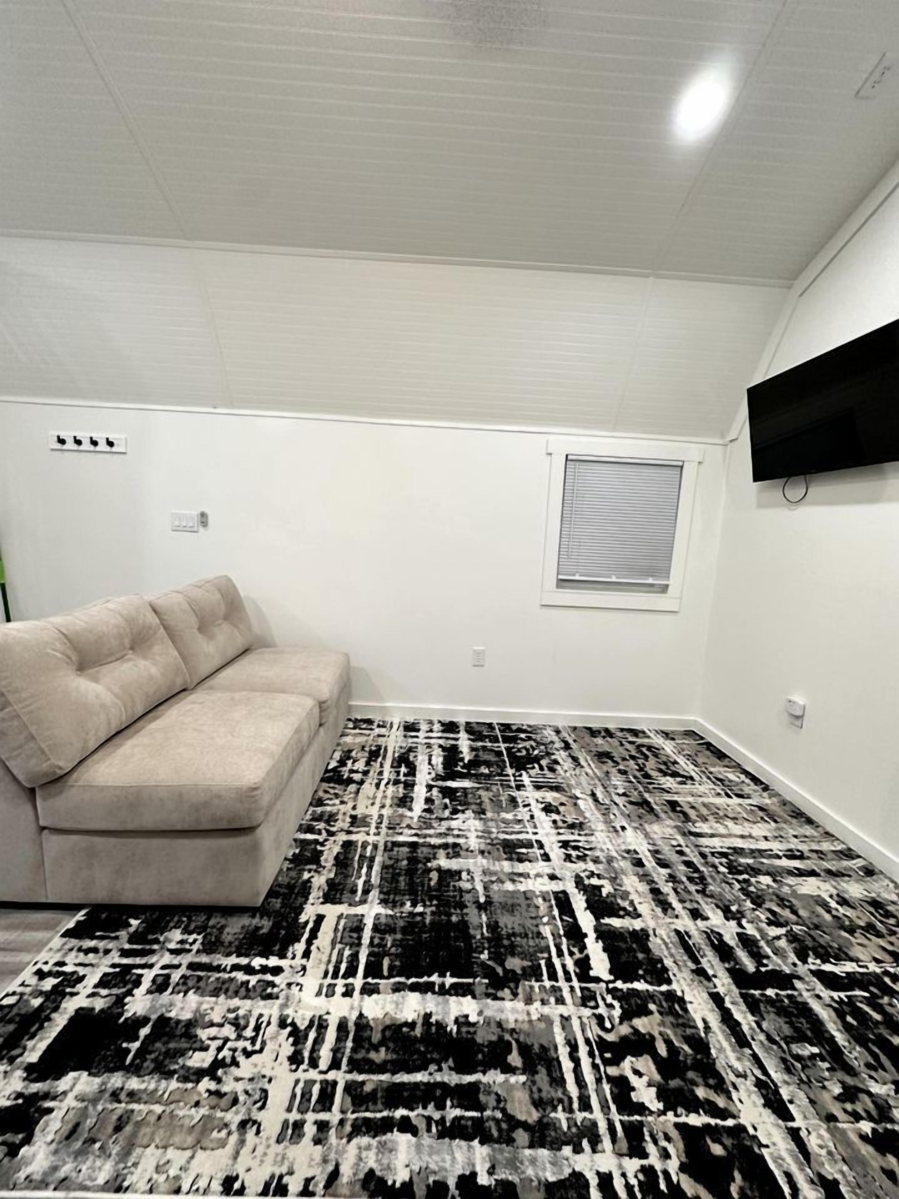 Une pièce avec un canapé beige, un tapis à motifs, une fenêtre avec des stores et un téléviseur fixé au mur.