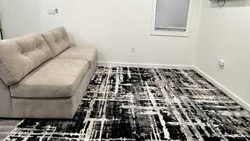 Une pièce avec un canapé beige, un tapis à motifs, une fenêtre avec des stores et un téléviseur fixé au mur.