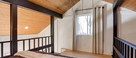 A bedroom with a sloped ceiling, a bed, a window with curtains, and a view of trees outside.