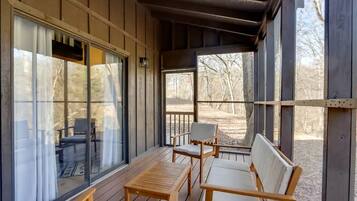 Un porche techado con muebles de madera, ventanas amplias y vistas a árboles.