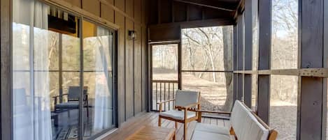 Un porche techado con muebles de madera, ventanas amplias y vistas a árboles.