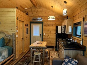 A cabin interior with a wooden interior, a dining table with stools, a kitchen area, and a bed with a headboard. - Scenic Creekside Escape with Deck, Fire Ring & Wildlife Views in Seward, Alaska (Seward)