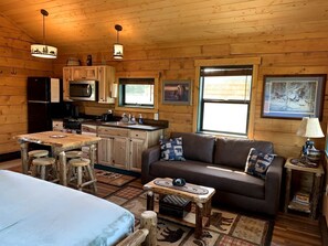 A cozy cabin interior with a wooden dining table, a sofa, and a kitchen area. - Scenic Creekside Escape with Deck, Fire Ring & Wildlife Views in Seward, Alaska (Seward)