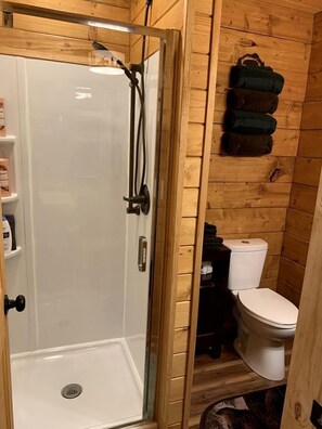 A wooden-paneled bathroom with a glass-enclosed shower, a toilet, and a shelf with toiletries. - Scenic Creekside Escape with Deck, Fire Ring & Wildlife Views in Seward, Alaska (Seward)