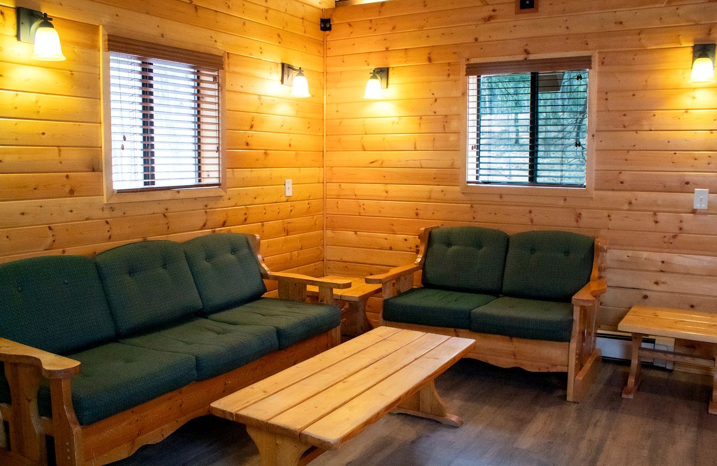 Interior de una cabaña de troncos con un sofá verde, dos sillones, una mesa de madera y dos ventanas con persianas.