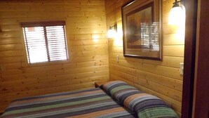 Een kamer met houten wanden, een bed met gestreept beddengoed, een raam met jaloezieën en een wandlamp.