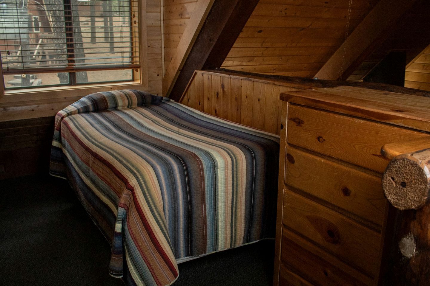 A small, cozy room with a sloped ceiling, a single bed with a striped blanket, a wooden dresser, and a window with blinds.