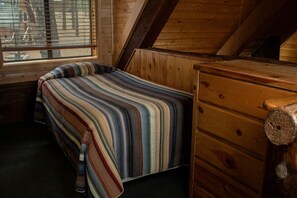 Una habitación pequeña y acogedora con techo inclinado, una cama individual con manta a rayas, un aparador de madera y una ventana con persianas.