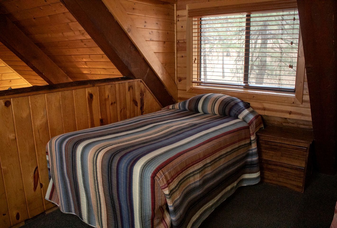 A small, cozy room with a sloped ceiling, a single bed with a striped bedspread, a window with blinds, and a wooden dresser.
