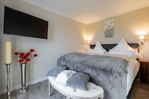 A bedroom with a large bed, bedside lamps, a flat-screen TV, a decorative vase with red flowers, and a fluffy grey blanket.