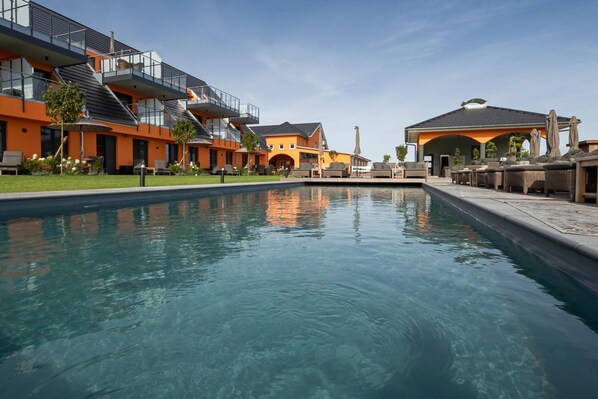 A swimming pool surrounded by modern buildings with balconies, outdoor seating, and a clear sky.