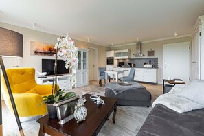 A modern living room with a yellow armchair, a grey sofa, a coffee table with a clock, and a kitchen in the background.