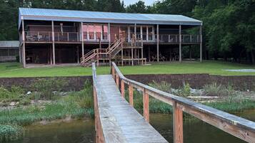 A wooden dock leading to a building with a metal roof, surrounded by greenery and a pond.