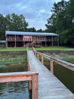 A wooden dock leading to a building with a metal roof, surrounded by greenery and a pond. - Lake Wateree Retreat,Private Dock, Fire Pit,Kayaks,Sunset glow views, sleeps 6 (Ridgeway)