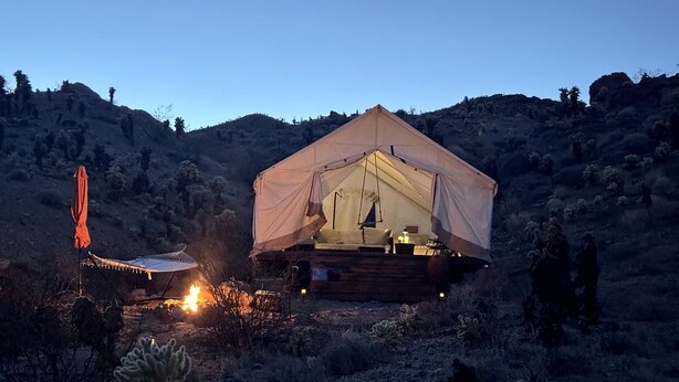 Eine Zelthütte mit Bett, kleinem Tisch und Stuhl, umgeben von Kakteen und Hügeln.
