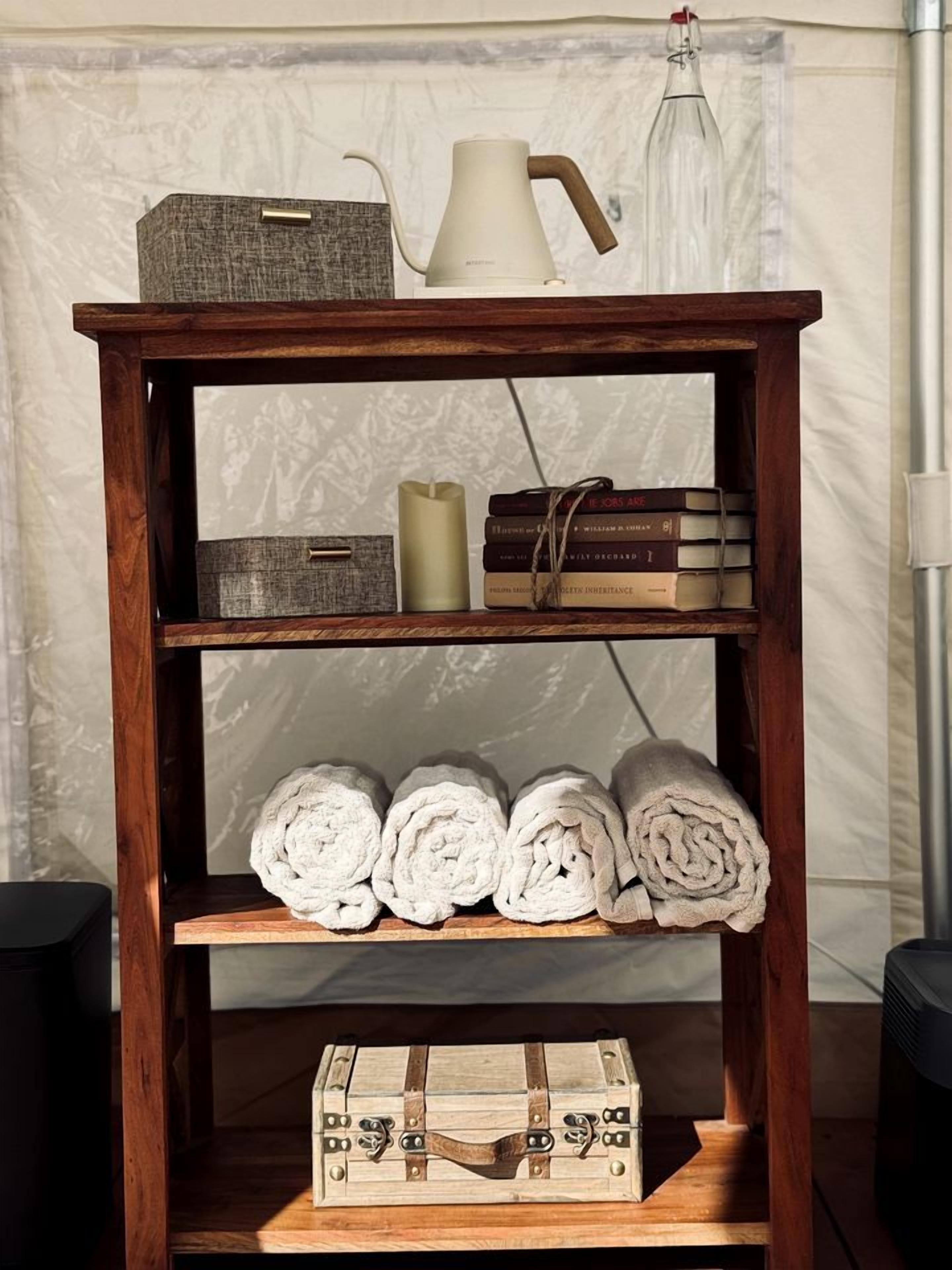 A wooden shelf with a white kettle, books, rolled towels, and a vintage suitcase.