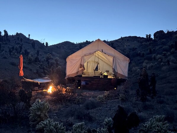 A tented accommodation with a bed, a small table, and a chair, surrounded by cacti and hills.