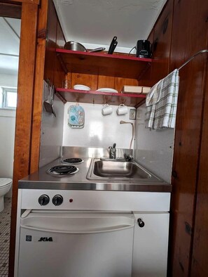 A compact kitchenette with a stainless steel stove, a sink, and a refrigerator. There are wooden cabinets above the stove and a small shelf with dishes. - Cozy Oceanfront Escape with Stunning Views in North Truro, Massachusetts (North Truro)
