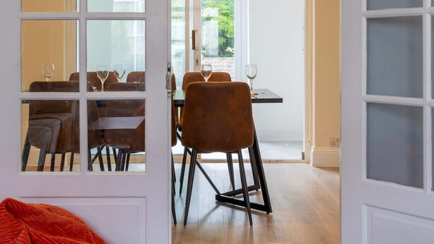 Une porte ouverte donne sur un coin repas où une table est dressée pour deux personnes, avec un fauteuil en forme de sac de haricots rouge et une vue sur la verdure à l’extérieur.