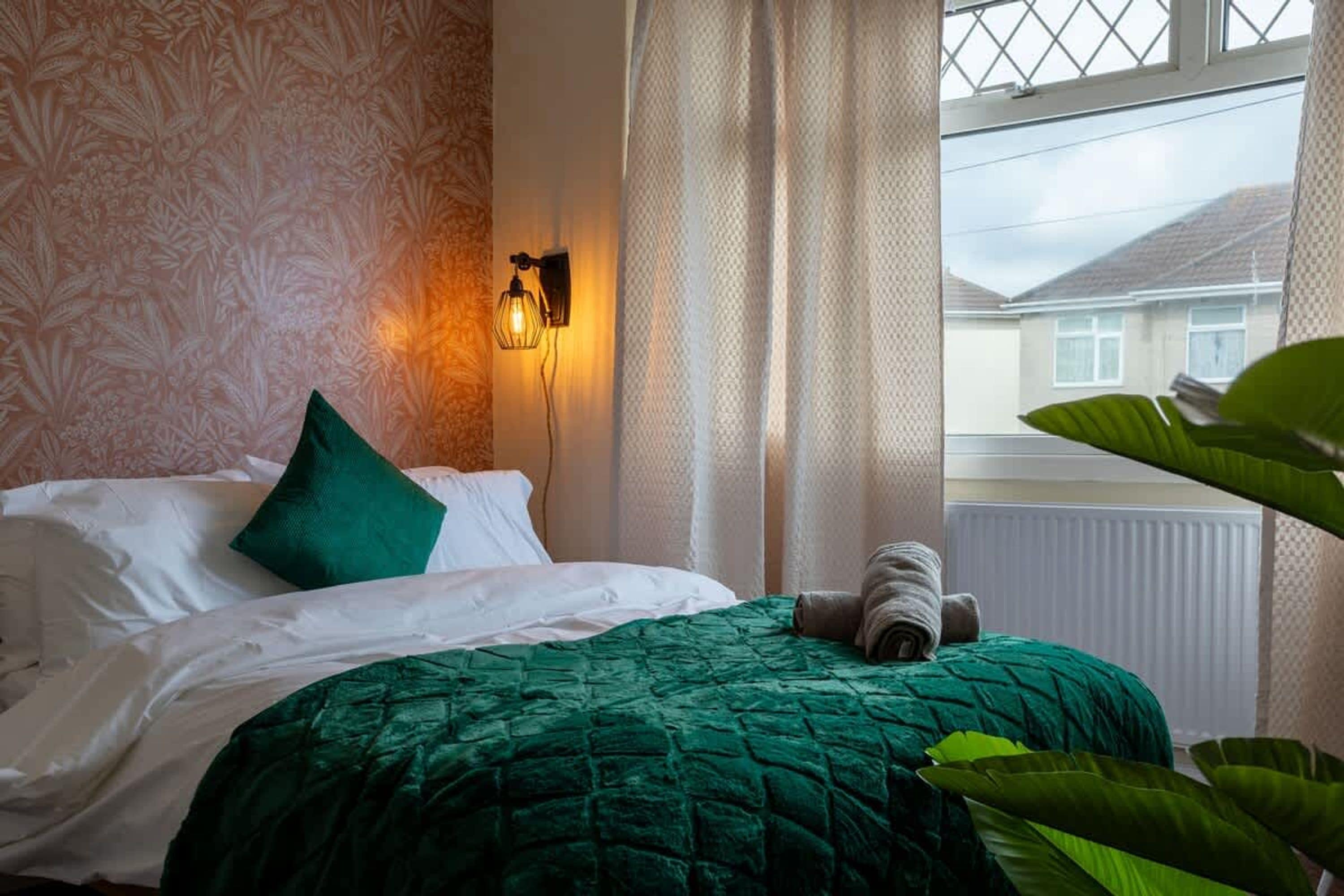 A bed with a green quilt, a green pillow, and a rolled towel. There is a wall-mounted light fixture and a window with curtains.