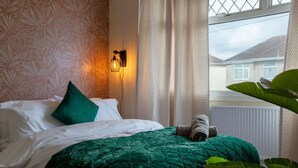 Un lit avec une couette verte, un oreiller vert et une serviette enroulée. Il y a un luminaire fixé au mur et une fenêtre avec des rideaux.