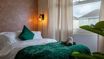 A bed with a green quilt, a green pillow, and a rolled towel. There is a wall-mounted light fixture and a window with curtains.