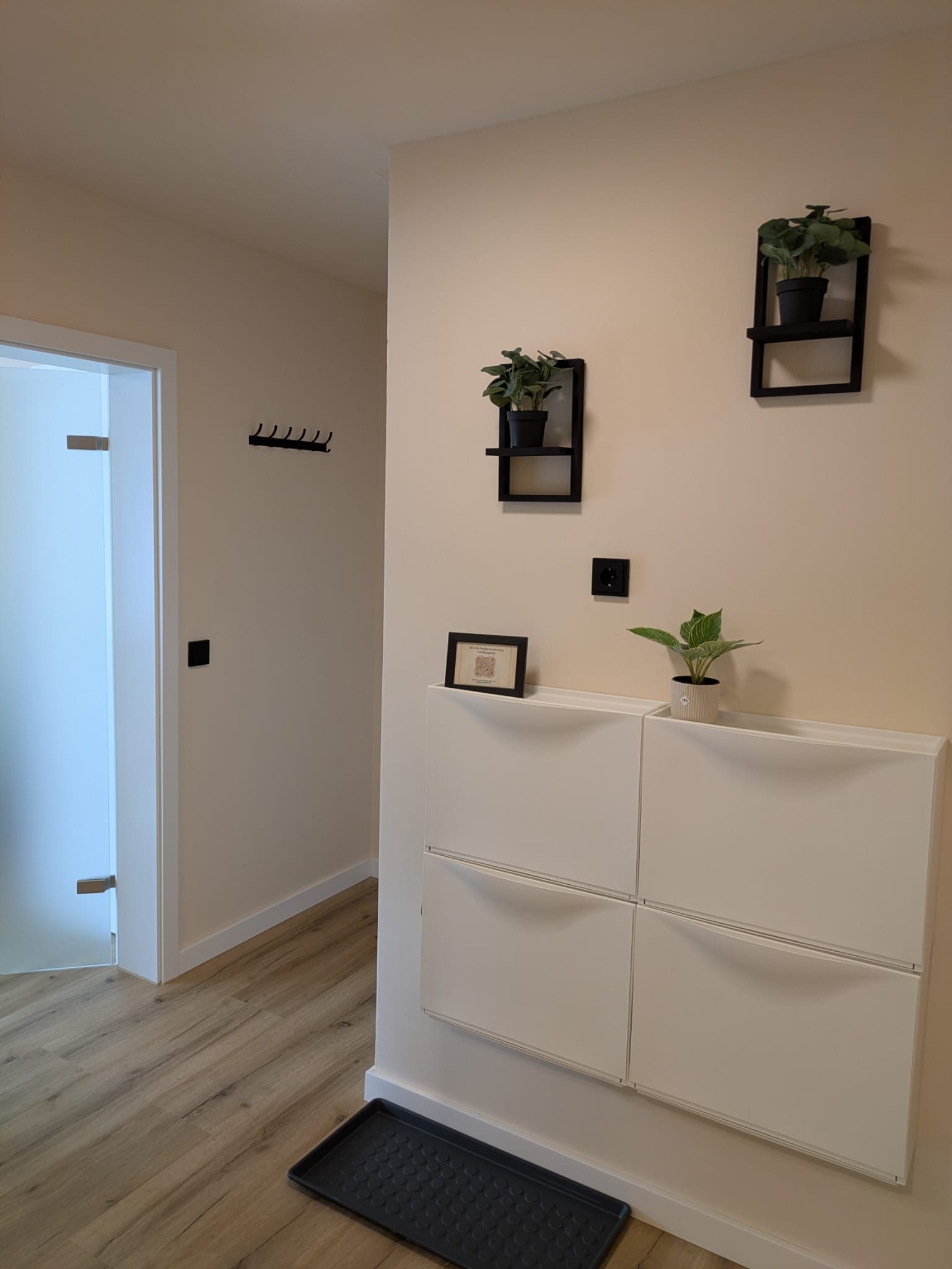 Ein Flur mit einer weißen Tür, einem Kleiderständer, einem an der Wand befestigten Regal mit einer Pflanze und einer gepolsterten Wandverkleidung in Weiß.