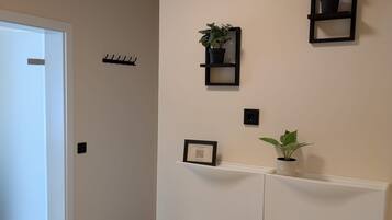 A hallway with a white door, a coat rack, a wall-mounted shelf with a plant, and a white padded wall unit.