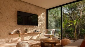 A modern living room with a stone wall, a flat-screen TV, a large window with a view of trees, and a cozy seating area with a coffee table and poufs. - Villa Lumila Rooftop Sunset & Jungle Views (Tulum)