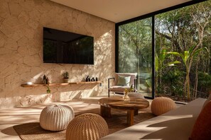 A modern living room with a stone wall, a flat-screen TV, a large window with a view of trees, and a cozy seating area with a coffee table and poufs. - Villa Lumila Rooftop Sunset & Jungle Views (Tulum)