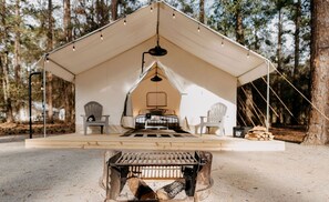 Ein Zelt mit Bett, Stühlen und einer Feuerstelle in einer Waldlandschaft.