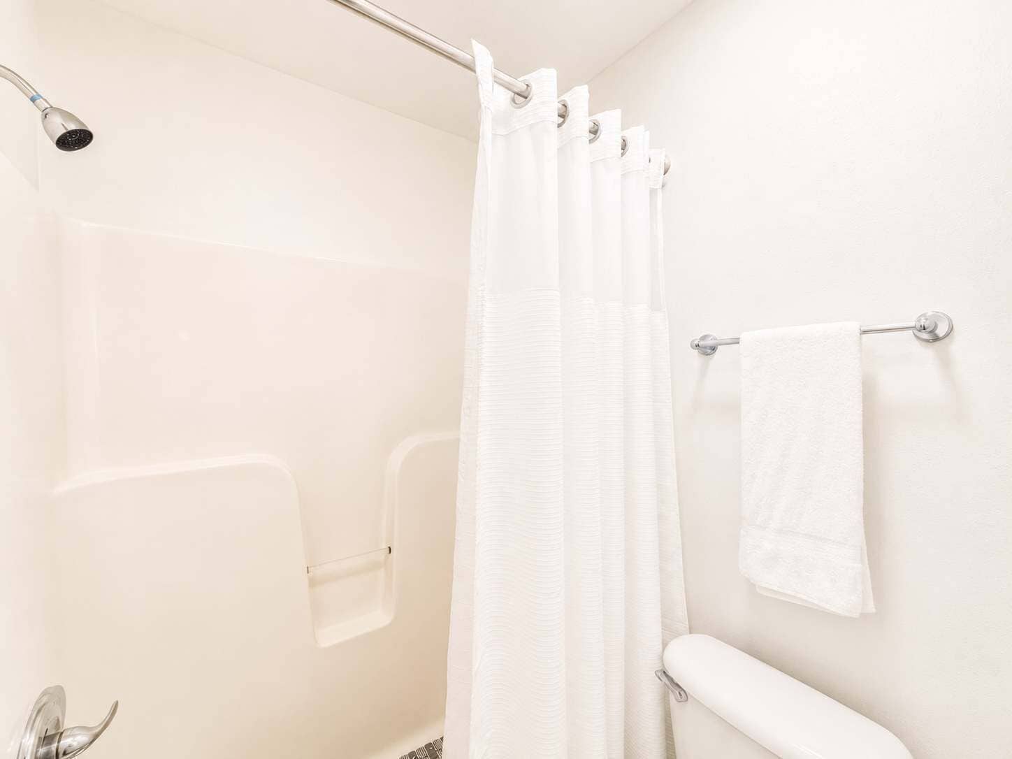 Un baño con ducha, cortina blanca, toallero y barra de apoyo.