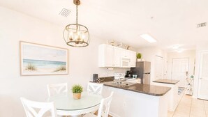 A modern kitchen with white cabinets, a central island, and a dining area with white chairs. - Grand Caribbean - 315 Newly Remodeled (Orange Beach)