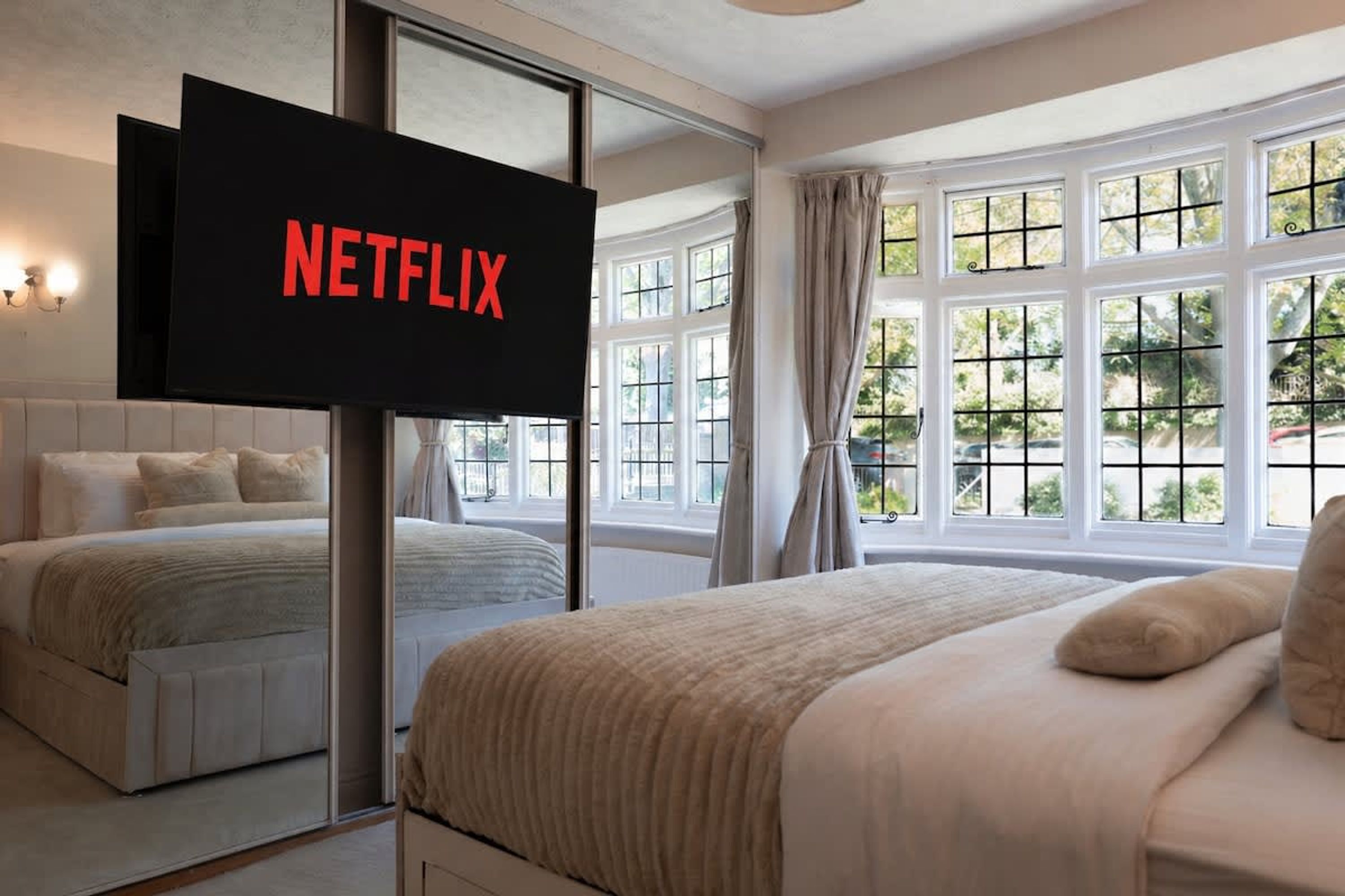 A bedroom with a large bed, a TV displaying 'NETFLIX,' and a view of the outdoors through large windows.
