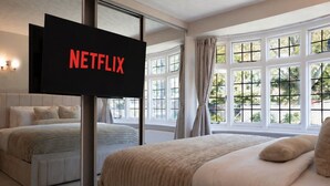Ein Schlafzimmer mit einem großen Bett, einem Fernseher, der 'NETFLIX' anzeigt, und einem Blick ins Freie durch große Fenster.