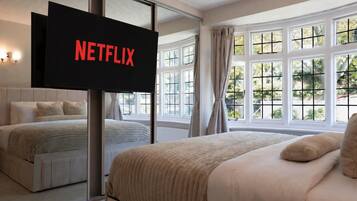 Et soveværelse med en stor seng, et fjernsyn der viser 'NETFLIX', og udsigt til det fri gennem store vinduer.