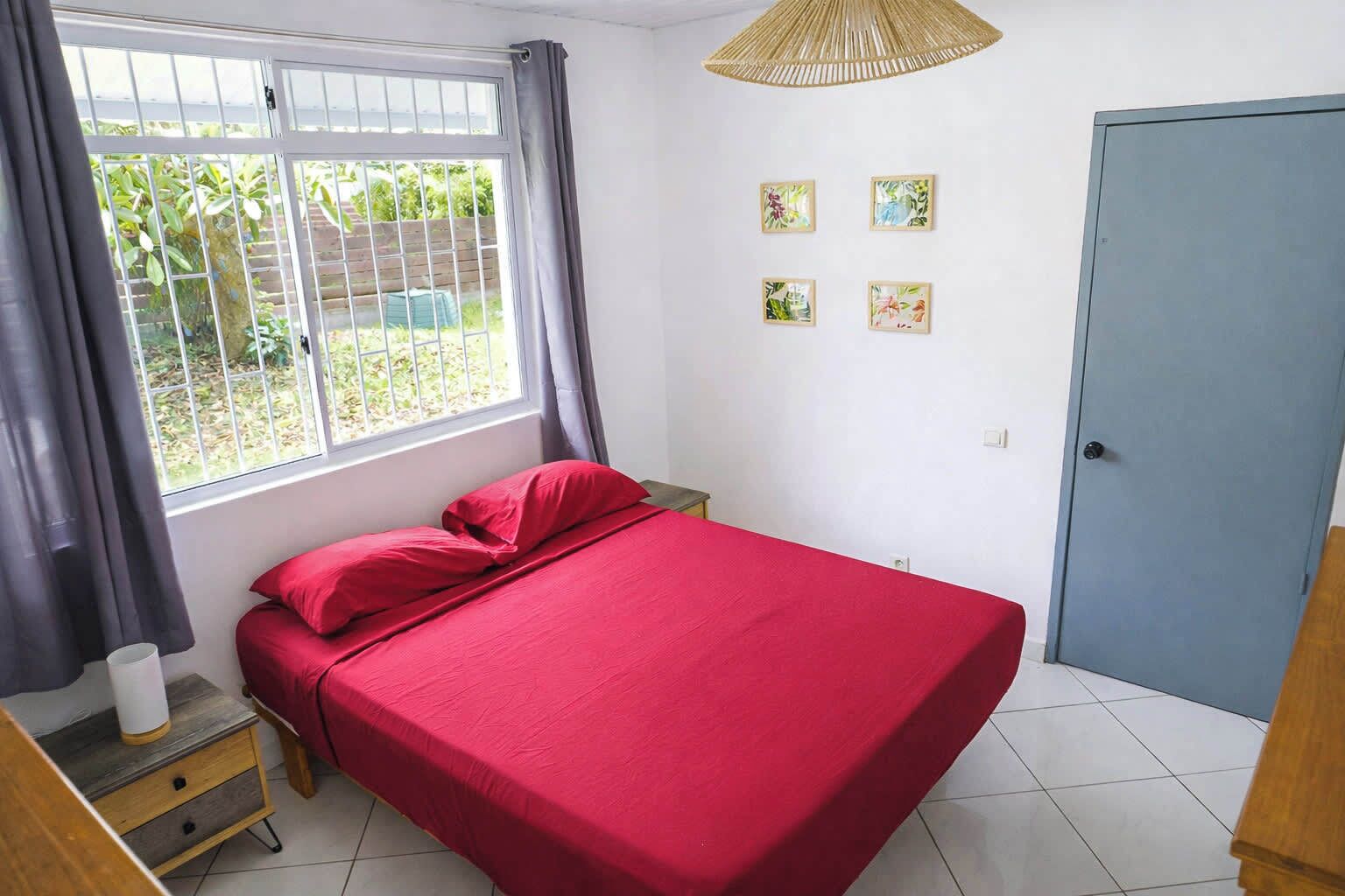 A bedroom with a bed, a window with curtains, a door, and a wall with four framed pictures.