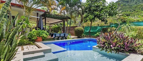 A swimming pool with steps leading into it, surrounded by tropical plants and a covered patio area.