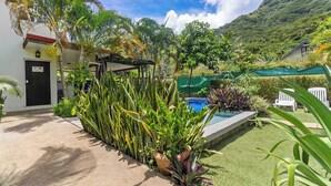 Piscina rodeada por vegetação exuberante, um edifício com uma porta identificada como 'Guesthouse' e montanhas ao fundo.