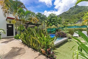 A pool surrounded by lush greenery, a building with a door labeled 'Guesthouse,' and a mountainous backdrop.