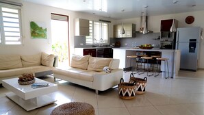 A modern living room with a sofa, coffee table, ottoman, and kitchen area with bar stools. - Dolce Vita Villa Moorea (Moorea-Maiao)