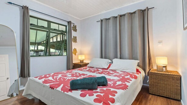 A hotel room with a bed, two bedside tables with lamps, a window with curtains, and a door. - Peaceful 2BR Garden Bungalow (Taiarapu-Est)